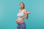 embarrassed young pretty white headed woman with natural makeup grimacing her face raising confusedly palm while posing blue background going be mom soon SKIN & HAIR - HOW PREGNANCY EFFECTS THEM?